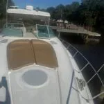 Front deck of the boat with two beige sun pads, round hatch, and railing, dock in the background.